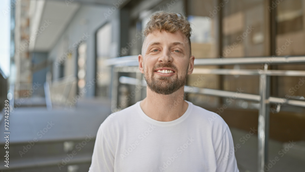 A handsome, bearded young man with blue eyes wearing a white shirt smiles warmly on an urban city street.
