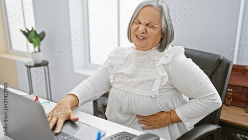Obraz na płótnie A mature woman grimacing in discomfort while working at her tidy office desk