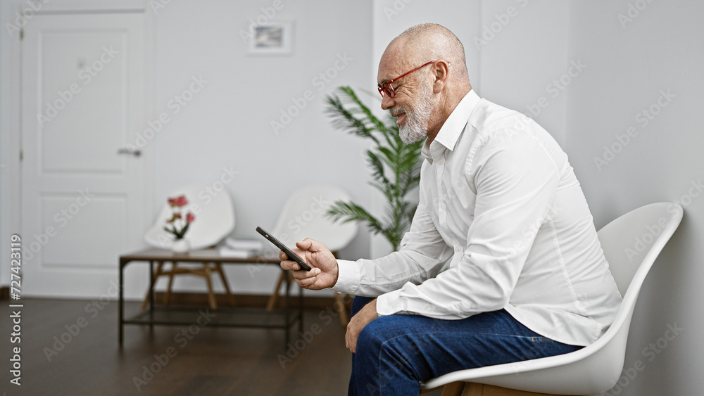 Bald man with beard in glasses using smartphone in modern waiting room interior