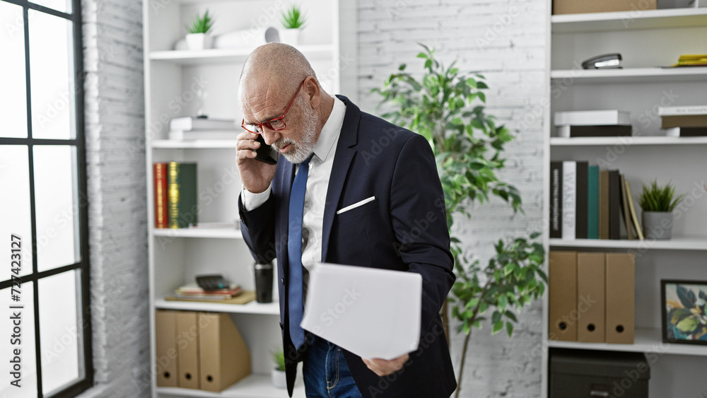 A mature bald man with a beard, wearing glasses, talks on a smartphone in a modern office.