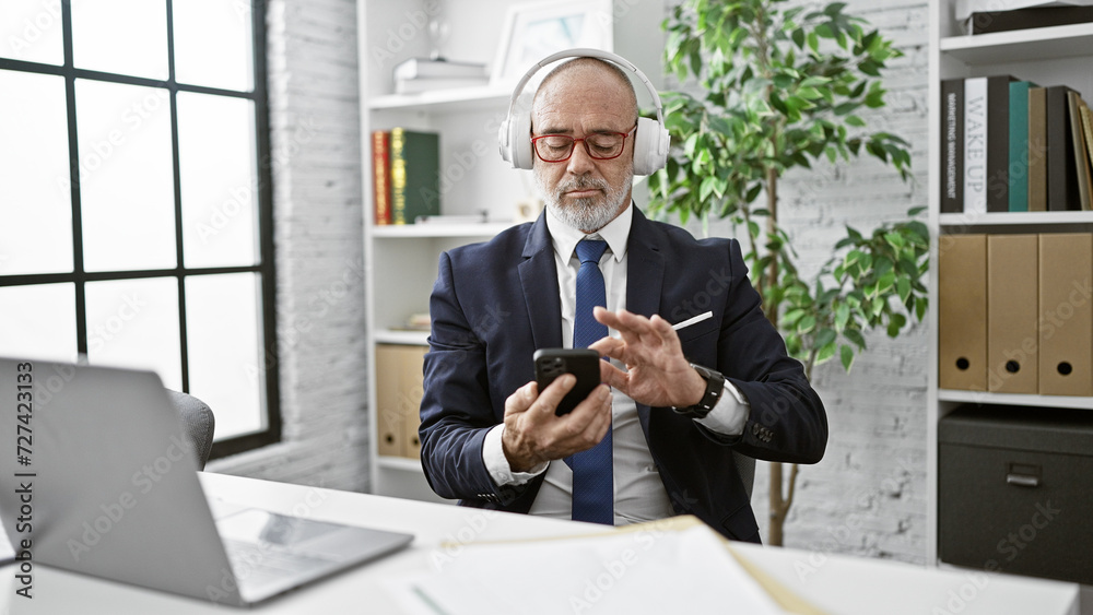 A senior businessman with headphones uses a smartphone in a modern office.