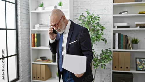 A mature bald man with a beard, wearing glasses, talks on a smartphone in a modern office.