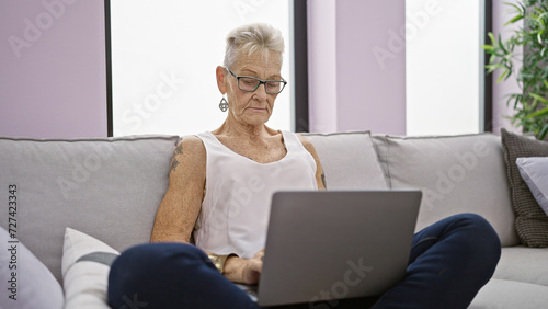 Focused elderly woman with grey hair and glasses, using laptop with concentration while sitting on sofa indoors at home, embodying mature technology use in the living room