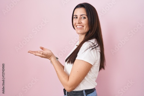 Young brunette woman standing over pink background pointing aside with hands open palms showing copy space, presenting advertisement smiling excited happy