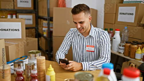 A smiling young man using a smartphone while volunteering in a donation center surrounded by boxes and food supplies.