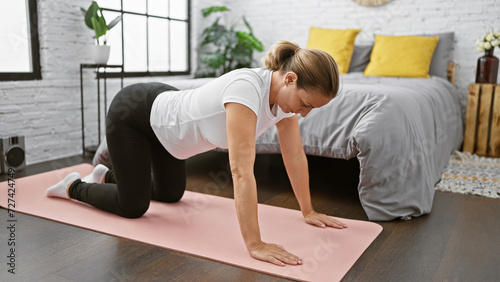 Attractive, young blonde woman in sportswear finds her morning balance by warming up with a calming back stretch on the bedroom floor next to her rumpled bed, smiling and stretching into the day. photo