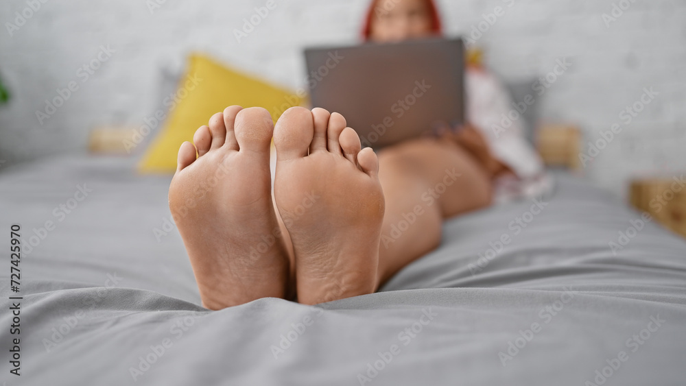 Fototapeta premium Relaxed morning moments, beautiful young redhead woman cozily lying on bedroom bed, comfortably wrapped in a blanket, engaging online with her laptop.