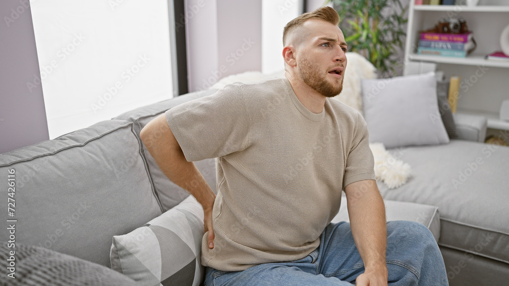 Young caucasian man with beard experiencing back pain at home, sitting on gray couch in a well-lit living room.