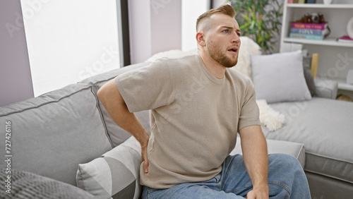 Young caucasian man with beard experiencing back pain at home, sitting on gray couch in a well-lit living room. © Krakenimages.com