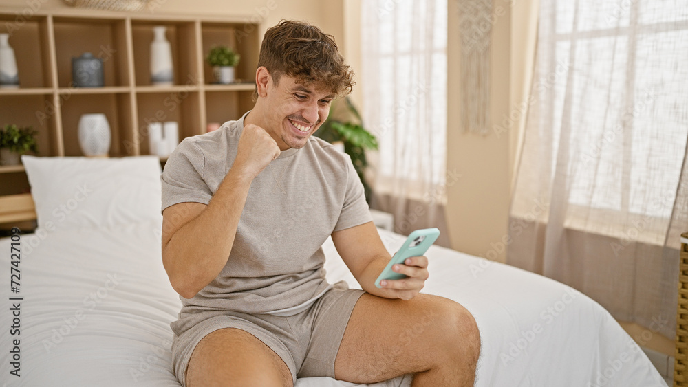 Relaxed young hispanic man sitting in cozy bedroom, all smiles as he celebrates his online win from the comfort of his bed, using smartphone Relaxed young hispanic man sitting in cozy bedroom, all smiles as he celebrates his online win from the comfort of his bed, using smartphone