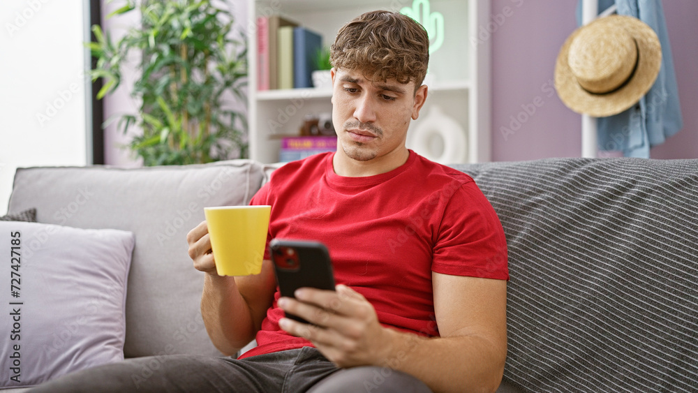 Handsome young hispanic man relaxing at home, concentrated on texting while drinking coffee on sofa