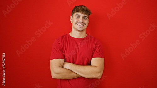 Cheery young hispanic man exuding confidence, standing tall with crossed arms, beaming a smile over an isolated red wall, radiating positivity, joy and infectious laughter