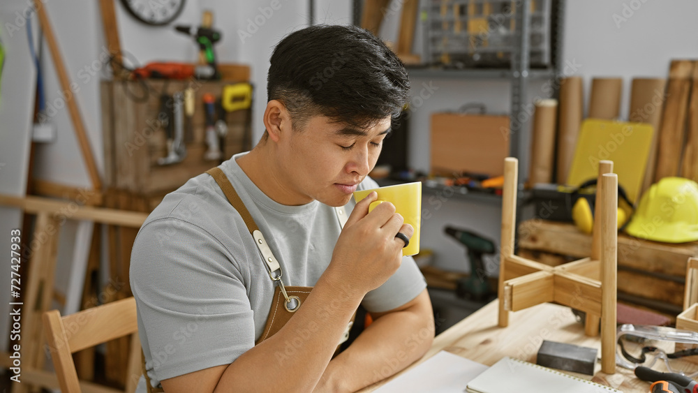 Young asian man enjoys a coffee break in a well-equipped carpentry workshop, surrounded by tools and wood.