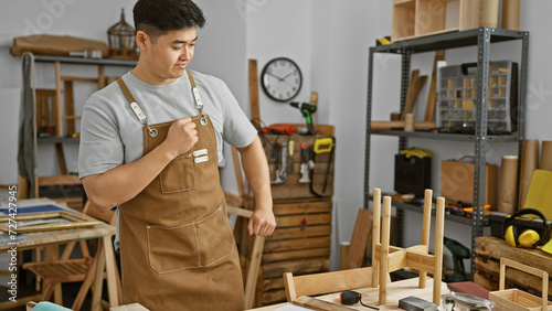 A young asian man in a workshop wearing a brown apron examining woodworking tools amidst various carpentry projects.