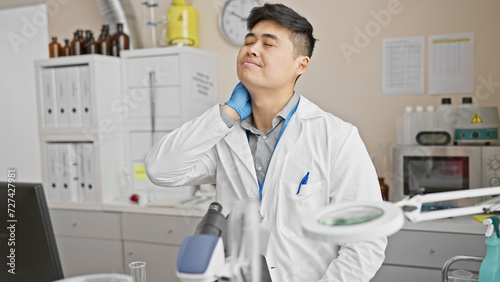 A young asian man in a white lab coat appears tired or stressed in a clinical laboratory setting.