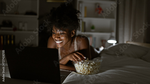 African american woman wearing lingerie watching movie on laptop eating popcorn at bedroom