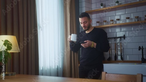 Smiling man standing in modern kitchen having morning coffee, holding cell phone scrolling news feed, checking emails, social media on smartphone photo