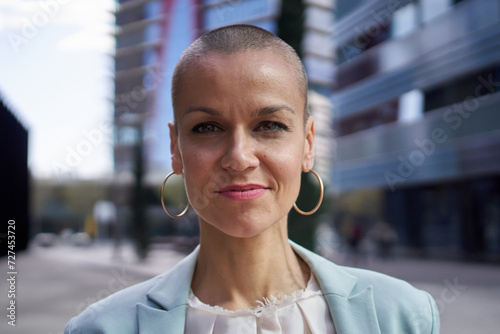 Close-up portrait of empowered middle-aged Caucasian business woman looking confident at camera. Formal attractive female posing elegant outdoors. Shaved head white person with successful expression photo
