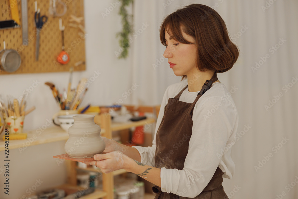 Professional potter wearing apron holding unfired clay vase while standing in pottery studio Professional potter wearing apron holding unfired clay vase while standing in pottery studio