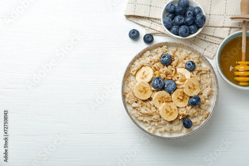 Tasty oatmeal with banana, blueberries, walnuts and honey served in bowl on white wooden table, flat lay. Space for text