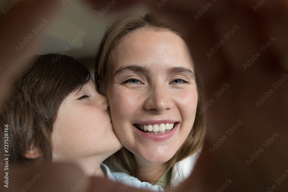 Close up shot, little loving son kiss mom cheek, making heart sign, look through fingers at camera, mother and kid demonstrate unconditional love to each other, enjoy tender moments. Happy motherhood