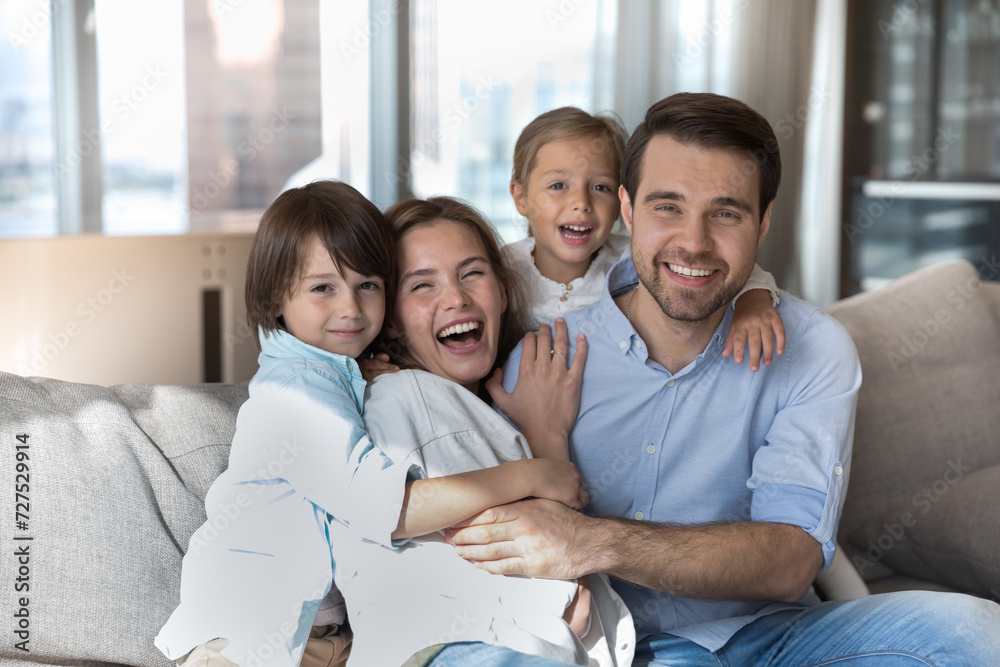 Cheerful homeowners couple and their adorable preschoolers children sit on sofa laughing, look at camera. Beautiful wellbeing family portrait, medical insurance cover, happy parenthood, ties and love