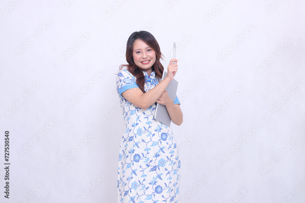 beautiful cheerful Indonesian Chinese woman looking at camera wearing modern Chinese blue dress hugging tablet and pointing pen top left isolated white background beautiful cheerful Indonesian Chinese woman looking at camera wearing modern Chinese blue dress hugging tablet and pointing pen top left isolated white background