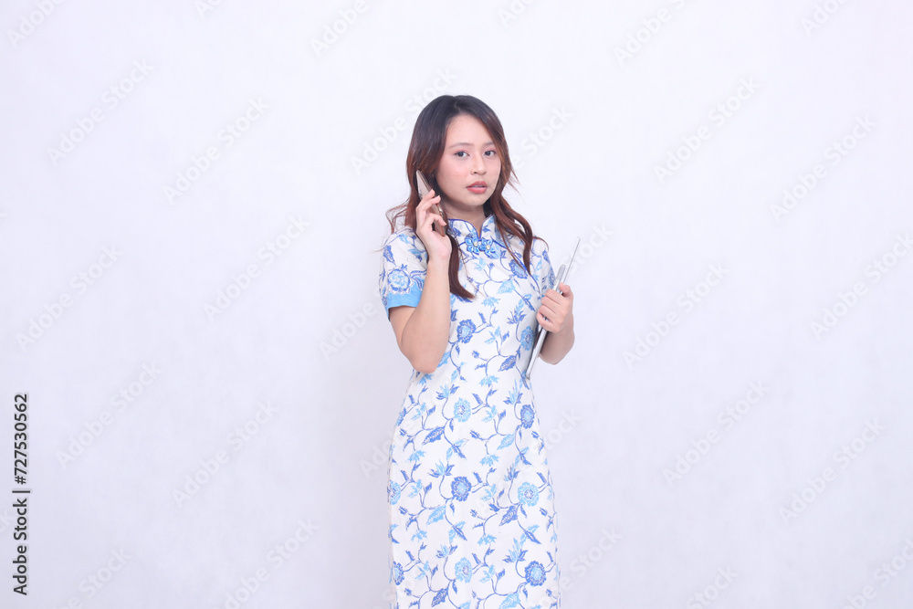 Beautiful Indonesian Chinese woman looking at the camera wearing a modern Chinese blue dress holding a tablet while making a phone call isolated on a white background