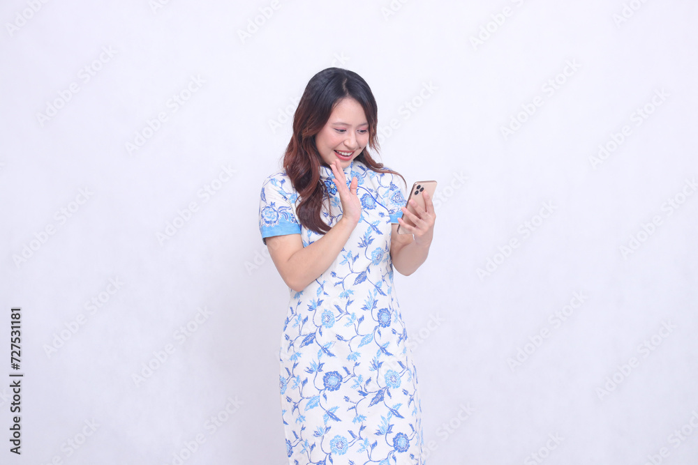 Young Asian Javanese Chinese woman smiles wearing a left-slanted Chinese blue dress while operating a gadget saying hello to communicating isolated on a white background Young Asian Javanese Chinese woman smiles wearing a left-slanted Chinese blue dress while operating a gadget saying hello to communicating isolated on a white background