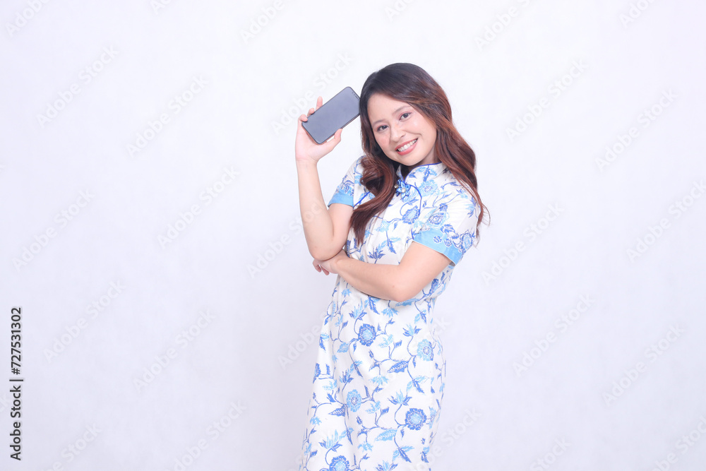 young Indonesian Javanese woman wearing blue traditional Chinese dress happy holding gadget showing tilted cell phone screen isolated white background young Indonesian Javanese woman wearing blue traditional Chinese dress happy holding gadget showing tilted cell phone screen isolated white background