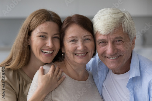 Positive old senior woman hugging mature husband and adult daughter with head close touches, looking at camera, smiling, laughing. Cheerful pretty woman and mature parents head shot portrait