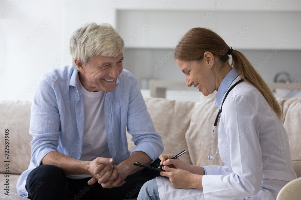 Obraz premium Positive elderly man consulting young doctor woman at home, speaking to medical specialist, smiling, laughing, feeling healthy. Practitioner visiting senior male patient in apartment, writing notes