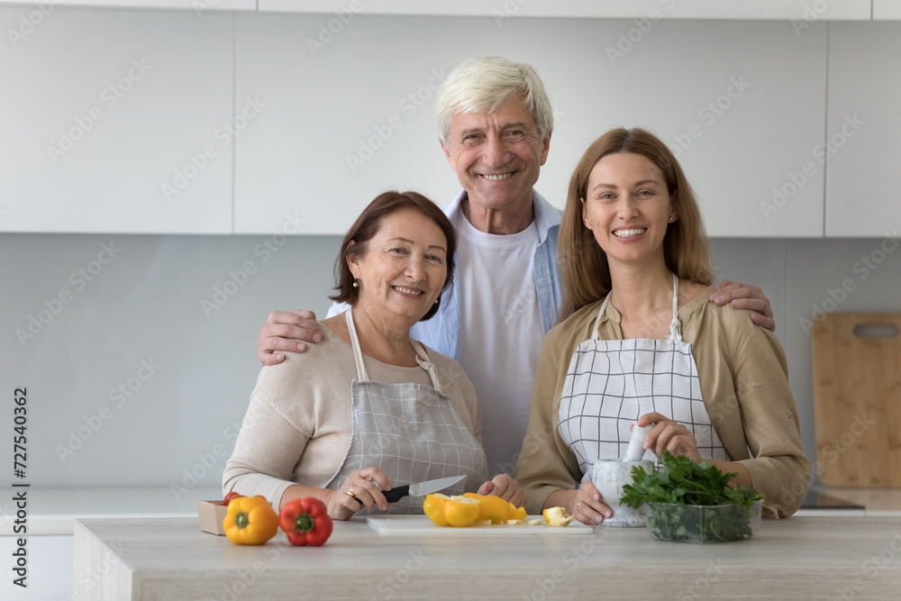 Happy mature old couple of parents and adult kid cooking family dinner in kitchen, chopping fresh vegetables for salad, looking at camera, enjoying leisure, holiday, culinary hobby. Front portrait