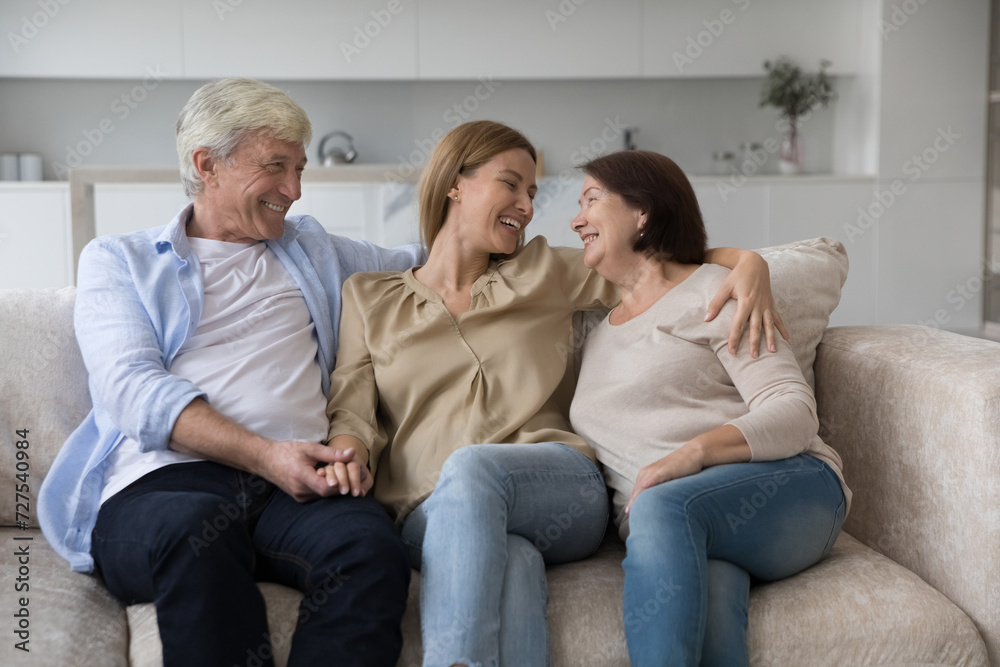 Positive caring senior parents and loving adult daughter woman enjoying leisure, closeness, sitting on couch at home together, hugging, smiling, discussing family news, laughing