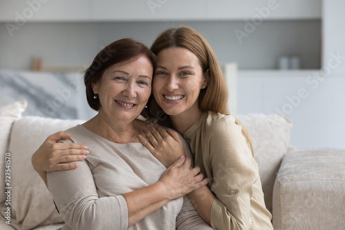Positive beautiful adult daughter child hugging older mom, touching shoulders with love, gratitude, care, support, visiting parent at home on mothers day, looking at camera, smiling, laughing