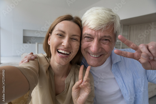 Funny family self portrait of cheerful senior dad and pretty adult daughter child holding device in hand, smiling at camera, laughing, taking home selfie making video call, showing victory fingers