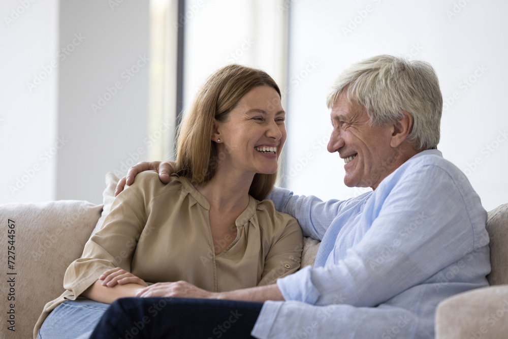 Cheerful adult daughter kid and happy senior grey haired dad talking, chatting on couch at home, smiling, laughing at joke, hugging, enjoying funny family leisure time together