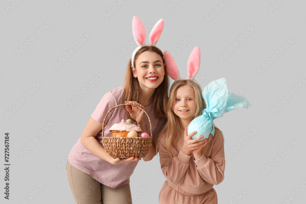 Cute little girl with gift egg and her mother in bunny ears holding basket with Easter cake on grey background