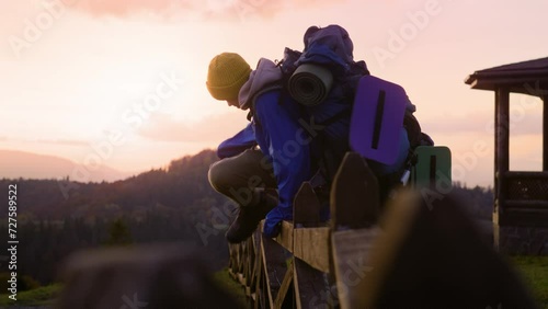 Multiethnic young tourists near wooden fence look at beautiful sunset and mountain landscapes enjoying nature during trip. Two teenage boys rest after hike or expedition in the mountains. Slow motion. photo