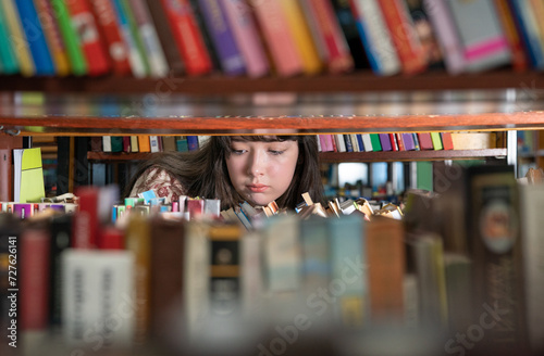 Girl standing between bookshelves in university library, picking up book from shelf, female student choosing textbook, looking for literature while preparing for exam. Education