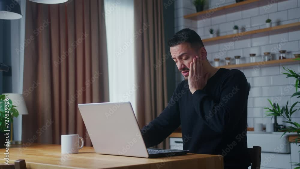 Tired man having pain from a headache or migraine rubbing his head while using laptop in the kitchen at home Tired man having pain from a headache or migraine rubbing his head while using laptop in the kitchen at home