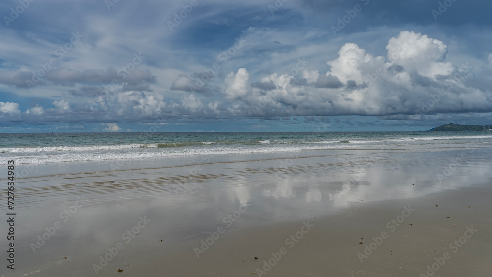 A serene seascape. The waves of the turquoise ocean roll towards the shore, foaming and spreading along the beach. Picturesque clouds in the blue sky.  Reflection on the wet smooth sand. Malaysia. 