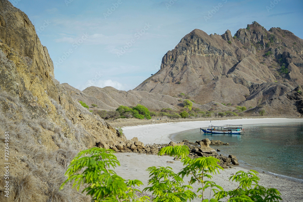 A beach scenery with semi circular white sand water front, a small boat tight to the shore, some green foliage in the foreground and around the beach, and majestic rock mountain and hills surrounding 