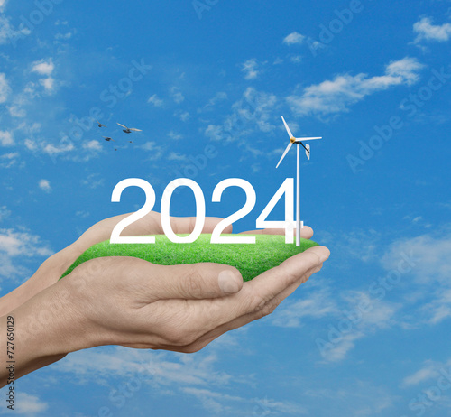 2024 white text with wind turbine on green grass field in hands over blue sky, white clouds and birds, Happy new year 2024 ecological cover concept