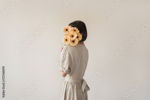 Beautiful young woman in neutral sandy linen dress holding gerber flowers bouquet over white wall. Aesthetic fashion background