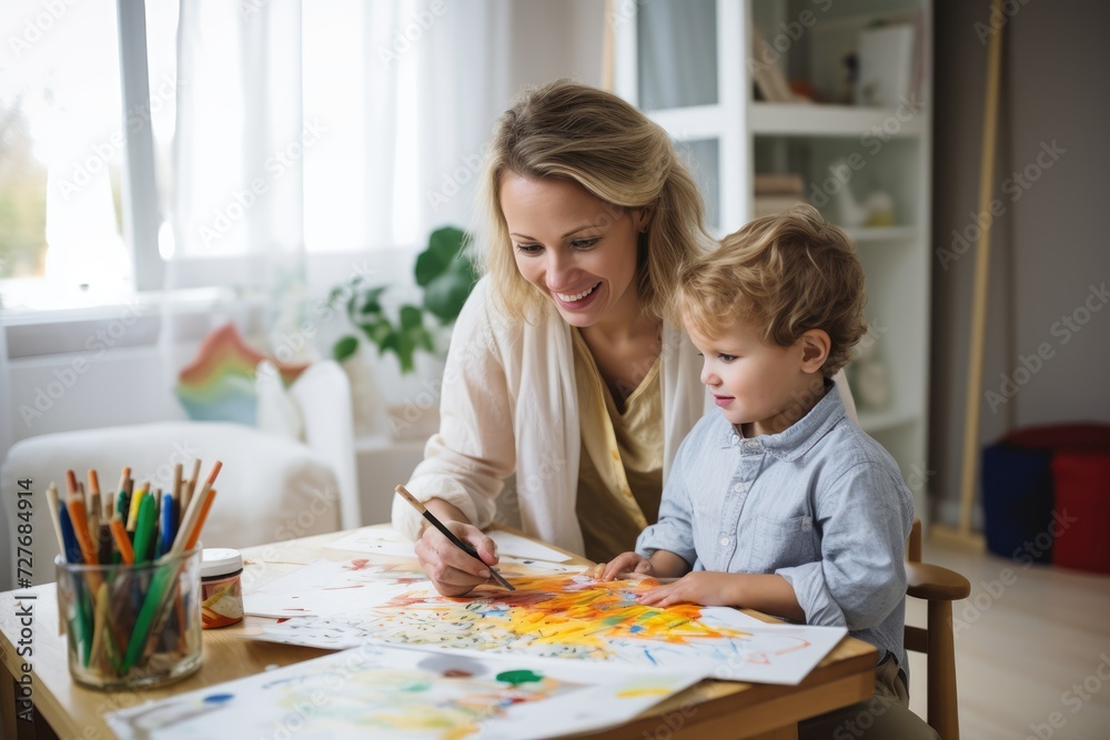 Fototapeta premium Beautiful woman and her cute little son are drawing and smiling while sitting at the table at home
