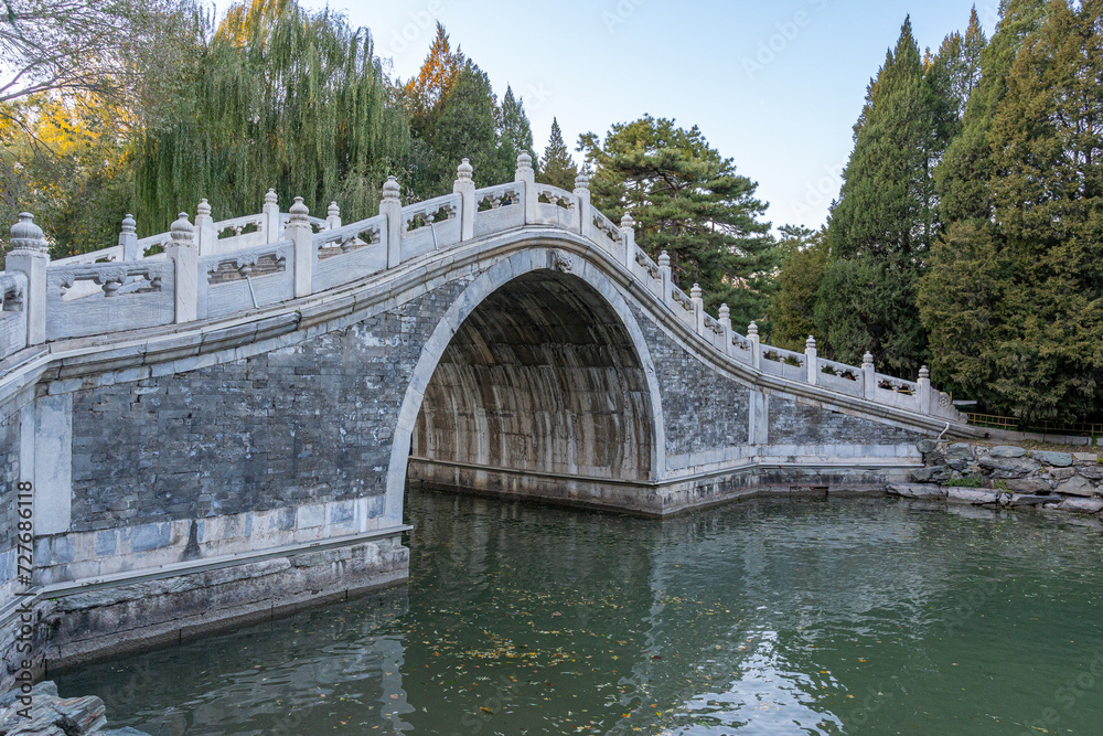 A beautiful white concrete bridge with a semi circular hollow center at the middle over a man made lake canal between trees under clear sky.