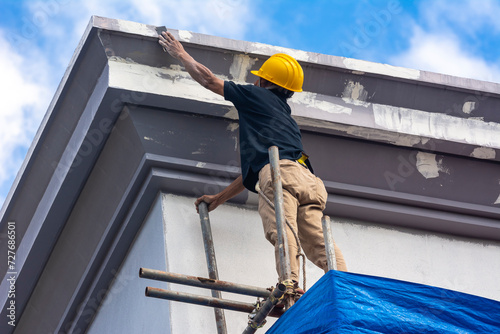 A skilled construction worker in a hard hat standing on top of scaffolding sanding the eaves and fascia of a 3 story house, in preparation for painting. Exterior renovation of a building facade.