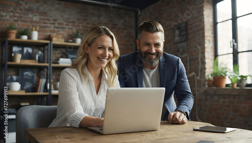 Smiling businesswoman and male creative director working together, talking about business strategy on a laptop computer in modern loft office.