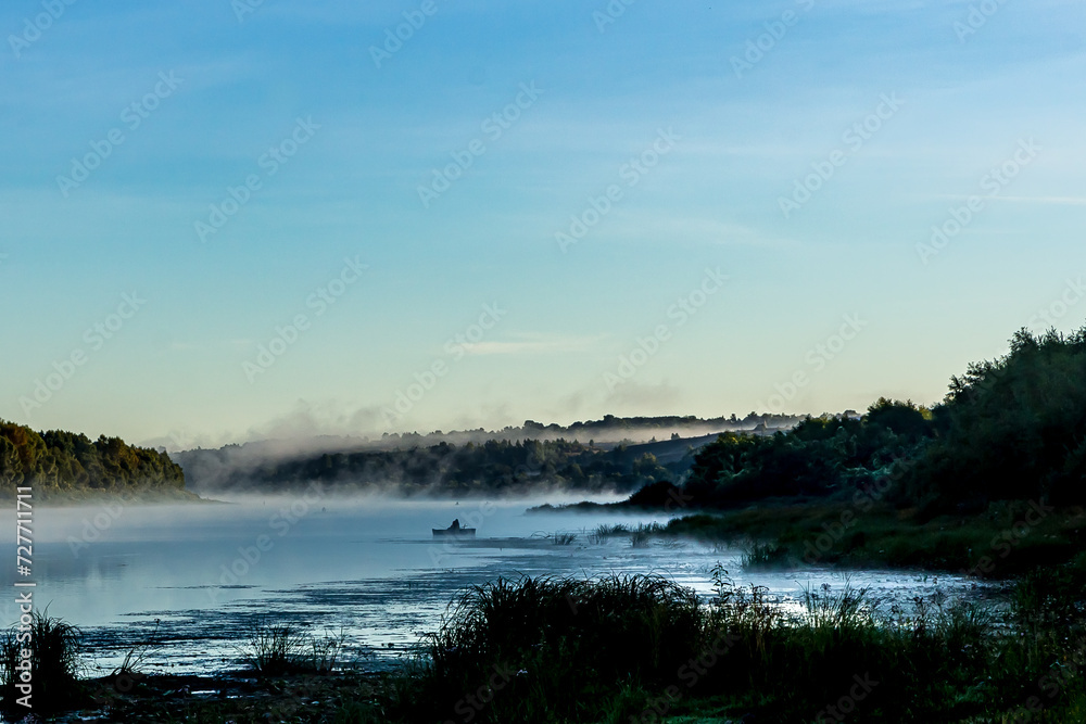 Fishermen on  boats early in the morning at dawn in the fog or mist at golden hour catch fish on the Oka River, Russia. Green grass and trees, a river bend against a clear light sky in summer.